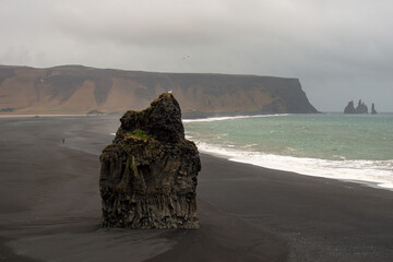 Dramatic sea stack on a black sand beach, with misty cliffs and crashing waves under an overcast sky.