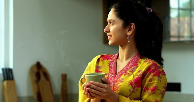 Indian Asian happy and pretty young girl in her twenties wearing salwar kameez and shawl, enjoying hot coffee or beverage in a modern kitchen with warm morning sunlight streaming in