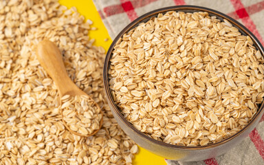 Rolled oats in a ceramic bowl with a wooden spoon on a yellow background and a checkered cloth. Healthy breakfast food concept.