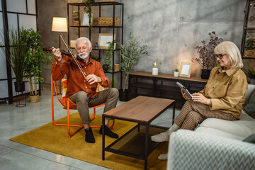 senior man play violin at home while his wife use digital tablet