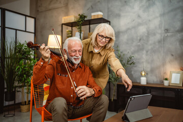 senior man learn to play violin at home on tablet and wife support him