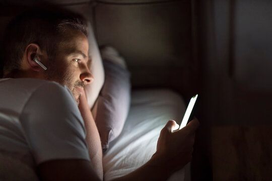 A sleepless man is lying in bed and doom scrolling on cellphone during bedtime.