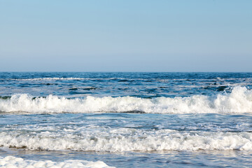 A serene beach scene with gentle waves rolling onto the sandy shore, white sea foam scattered along the water's edge, and a clear blue sky overhead