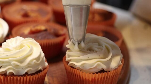 Cupcakes in orange liners are decorated with white frosting using a piping bag. The focus is on the intricate swirls, with undecorated cupcakes in the background.