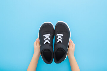 Little child boy hands holding and showing dark black sport shoes with white shoelaces on light blue table background. Pastel color. Footwear for children. Point of view shot. Top down view.