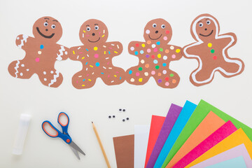 Brown gingerbread man shapes created from paper. Scissors, glue stick, pencil and colorful application paper on light gray table background. Making decoration elements. Closeup. Top down view.