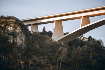 A modern concrete bridge spanning a lush green hillside with rocky formations and trees, illuminated by soft sunlight, showing the Ponte do Infante in Porto, Portugal
