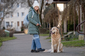Small Girl In Green Jacket Walks With Golden Retriever On Street In Early Spring, Showcasing A Child And Dog Bond