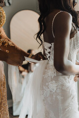 Mother adjusting and tying the intricate lace detailing of a bridal gown, capturing a tender...