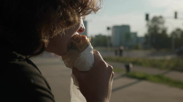 A young man bites off a piece of shawarma, watching city traffic pass by on the street. The guy eats a doner kebab with meat and vegetables wrapped in pita bread and takeaway wrapping paper.