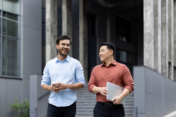 Two men walking and chatting outside an office building, colleagues and business partners taking a...