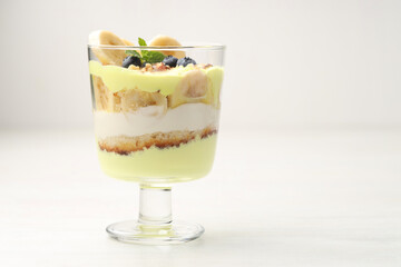 Tasty trifle dessert. Banana, blueberries, sponge cake and whipped cream in glass on white wooden table, space for text