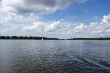 A scenic view of a calm river with sailboats gliding peacefully under a blue sky filled with fluffy clouds, perfect for nature lovers.