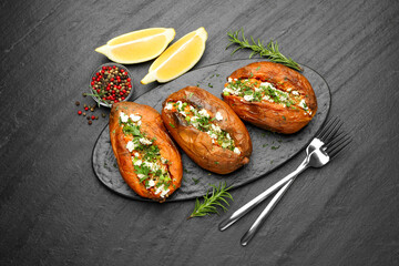 Tasty baked sweet potato with feta cheese, lemon, peppercorns and herbs on dark textured table, flat lay