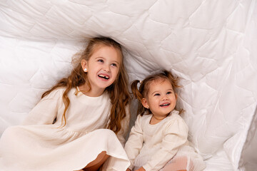 Children sisters in bed under the blanket having fun, smiling. Happy little girl and her newborn baby sister hiding under the white blanket. Childhood concept. Copy space. High quality photo