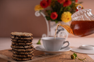 photo A Serene Tea Time Moment with a Glass Teapot Pouring Aromatic Tea into a Cup Surrounded by Delicious Cookies and Beautiful Floral Arrangement for a Cozy Afternoon