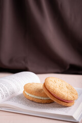 photo Delicious sandwich cookies with cream filling displayed on open book in front of luxurious brown fabric backdrop for cozy reading experience and sweet indulgence