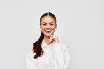 Smiling woman with long brown hair, wearing a white shirt against a plain light gray background, expressing confidence and happiness with a friendly gesture
