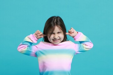 Annoyed little girl covering her ears from loud sound on light blue background