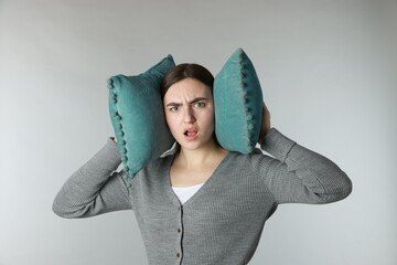 Distressed woman covering her ears with pillows from loud noise on light grey background