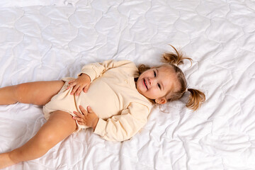 Naklejka premium Happy baby in bed. Cute little newborn girl in bodysuit lying on bed in bedroom. Infant baby resting, playing on white blanket at home. Motherhood and childcare concept