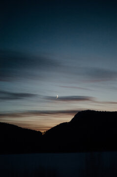 Moonlit crescent over silhouetted mountain ridge under a serene teal sky with wispy clouds during a winter evening..Europe, Norway, 02.01.2025  [The photographer confirms the authenticity of the photo