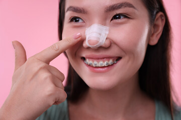 Woman with medical bandage on her nose after plastic surgery operation against pink background,...
