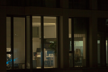 A modern office space with large, glowing windows is captured from the outside during the evening. The brightly lit interior stands out against the dark surroundings.