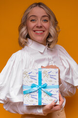 A smiling woman in a white blouse holds a floral-patterned gift box with a blue ribbon, set against...