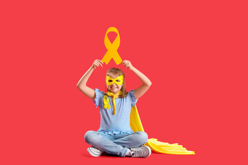 Little girl dressed as superhero with gold ribbon sitting on red background. Childhood cancer awareness concept