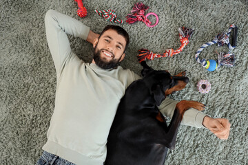 Young man with Dobermann dog and toys lying on carpet, top view