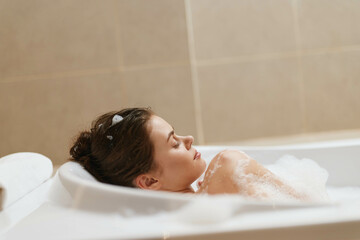 Relaxed young woman enjoying a soothing bubble bath in a modern bathroom, surrounded by soft light and calming colors for a peaceful atmosphere.