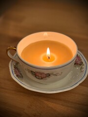 A homemade beeswax candle inside of an antique teacup and saucer. A perfect way to use old damaged heirloom china pieces. The candle glows on a wooden tabletop.