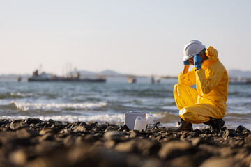 A person in a yellow protective hazmat suit and white helmet sits on a rocky shoreline, adjusting...