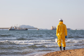 A person in a yellow hazmat suit and white helmet walking on a rocky shore, holding a water testing kit, conducting water quality checks. The background features the sea and distant boats © kamonrat