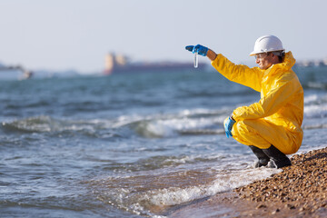 A person wearing a yellow protective hazmat suit and white helmet kneeling on a sandy shore,...