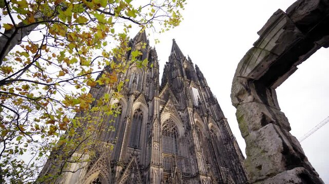 A majestic view of the Cologne Cathedral's twin spires framed by autumn leaves and ancient stonework, showcasing its intricate Gothic architecture under a cloudy sky