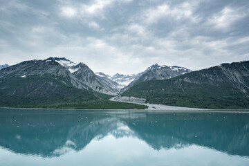 Fototapeta premium Beautiful snowy mountain peaks rising up from the shoreline of a calm body of water with a stunning reflection below and a cloudy sky overhead. 