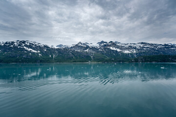 Beautiful snowy mountain peaks rising up from the shoreline with ripples in the blue water below and a cloudy sky overhead
