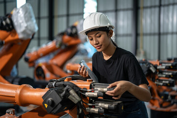 Female engineer wearing a white safety helmet and black shirt, standing in an industrial factory, analyzing orange robotic arms while taking notes on a clipboard. 