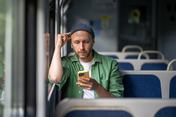 Man absorbed in music and podcasts through earbuds, traveling to work on train with smartphone in...