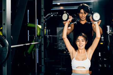 Sporty man and woman exercising with dumbbells, working on their muscle definition and wellbeing under the supervision of their coach, technique of exercise in gym dark gym background