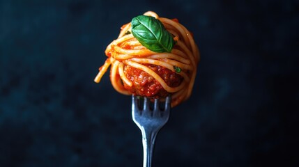 Spaghetti and meatball garnished with fresh basil on a silver fork against a dark background.