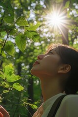 A woman in a forest, looking up at the sunlight filtering through the trees. She is wearing a light pink shirt and appears to be smiling.