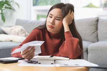 Business financial concept, stressed asian young woman hand checking list with bill for calculate accounting expense credit card payment on due deadline, trying to find money to pay debt on table.