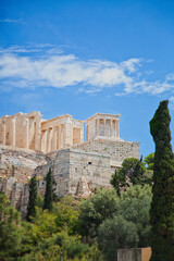 Fototapeta premium Ancient ruins of the Acropolis in Athens, Greece, stand majestically against a bright blue sky with scattered clouds, framed by lush green trees