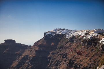 Stunning view of Santorini's white-washed buildings perched dramatically on volcanic cliffs overlooking the Aegean Sea. Traditional Greek architecture against rugged landscape and clear blue sky