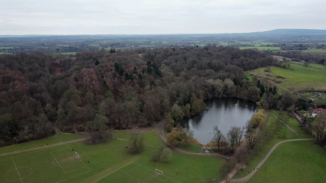 Stunning parallax aerial footage of Reigate Priory Lake, surrounded by lush woodland and open parkland. Features walking paths, football pitches, and serene countryside views under a cloudy sky.