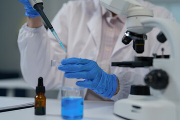 Fototapeta premium Close-up of scientist wearing blue gloves using pipette to drop blue liquid into test tube while conducting experiment in lab with microscope and other equipment