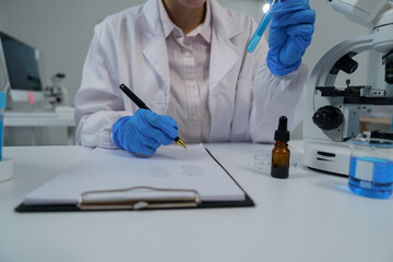 Scientist wearing blue gloves writing notes on a clipboard while holding a test tube containing...